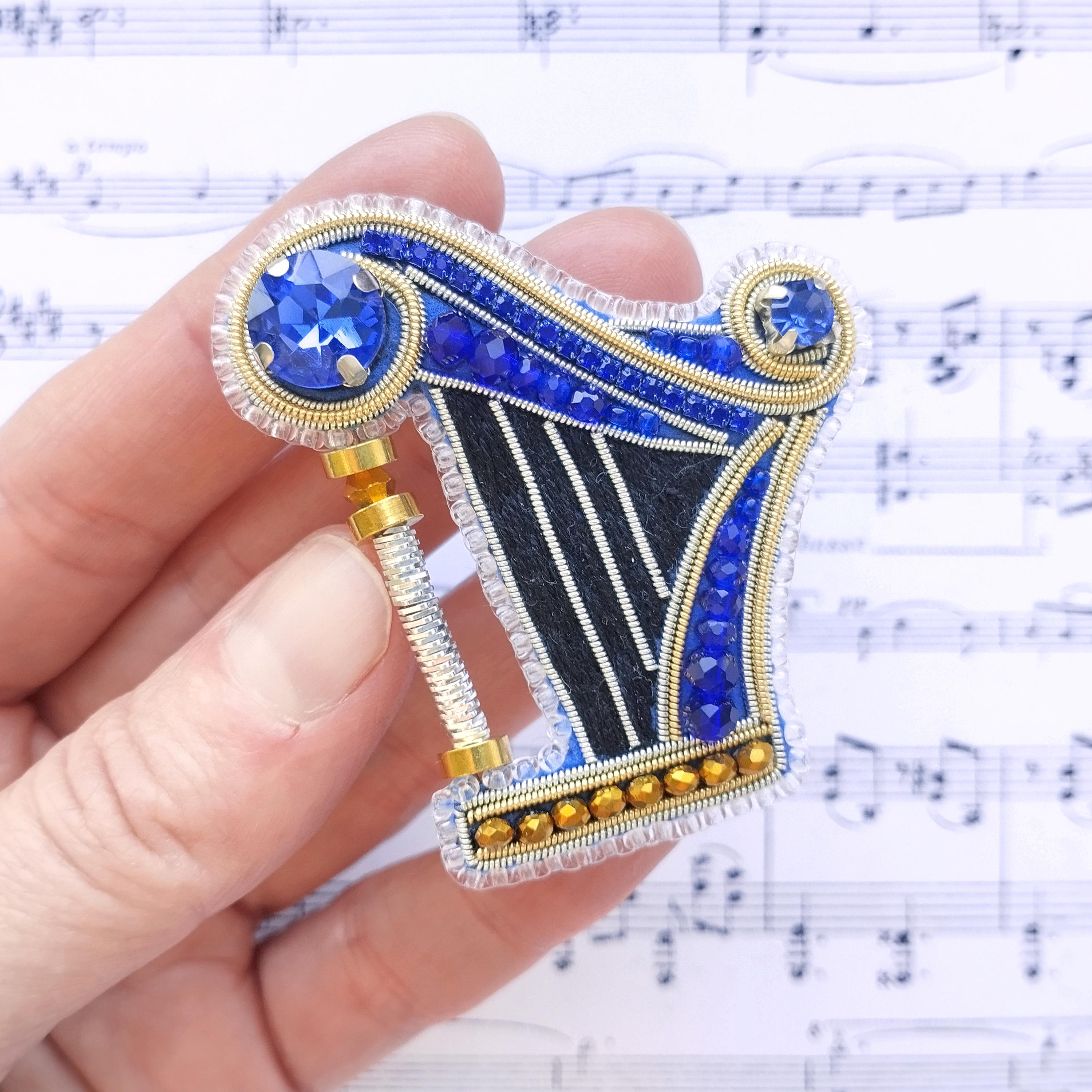 A hand holding a decorative piece with blue and gold elements, including a crystal-like gemstone and a metallic frame, against a background of sheet music.